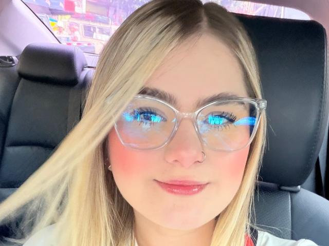 AlannaFoxy with blonde hair, blue eyes reflected in clear glasses, pink blushed cheeks, nose ring, and glossy pink lips, sitting in a car.