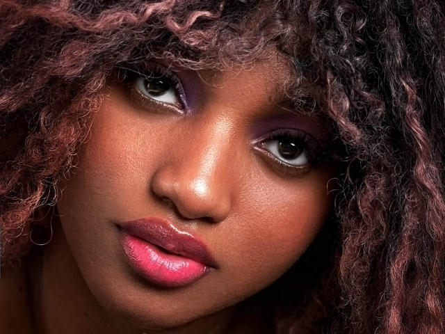 EmmaParisi's face with dark brown skin, pink glossy lips, purple eyeshadow, curly red-brown hair, and brown eyes looking slightly upward. Her smooth skin and full lips are prominent.