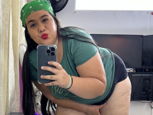AndreaaX69 with red lipstick, green bandana, green shirt, black shorts, bending over, showing her big ass and thick thighs, taking a selfie with a green phone.
