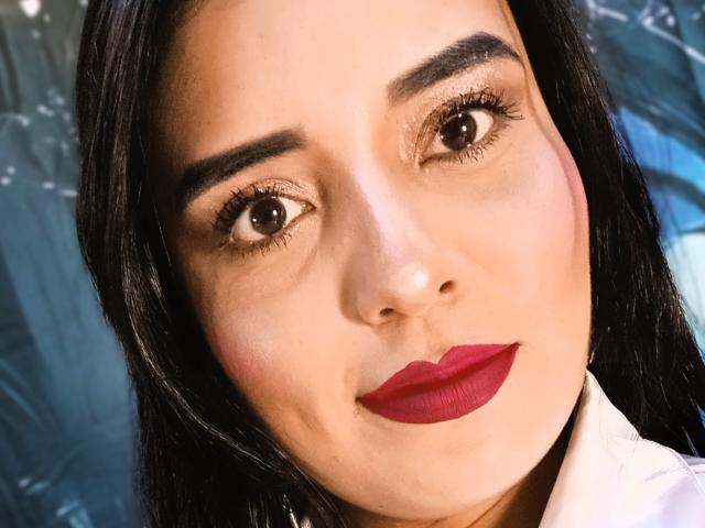 MelizaQueen's face with dark brown eyes, thick eyebrows, red lipstick, and black hair, looking directly at the camera. She has a light skin tone and a white shirt.