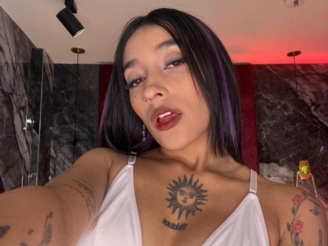 SalomeMille met zwart en paars haar, een witte tanktop, tatoeages, een zonnetattoo op de borst en rode lippenstift, met haar tong uit, kijkend naar de camera, in een slecht verlichte kamer..