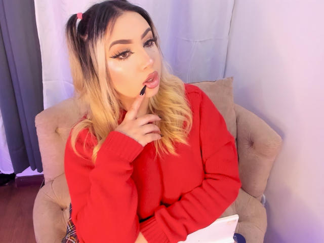 MelissaLust with blonde hair in pigtails, wearing a red sweater, biting her finger, sitting on a beige chair, looking at the camera, holding a white paper.