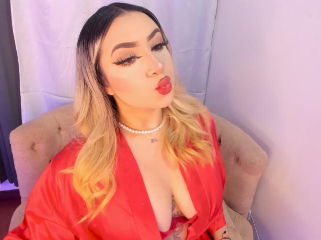 MelissaLust with blonde hair, red lipstick, and thick eyelashes, pouting, wearing a red silk robe, revealing her big tits, and a pearl necklace, sitting on a beige chair.