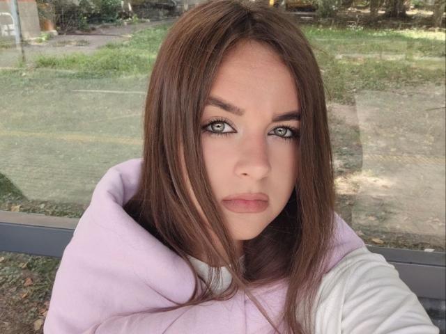 RozmaryKet has green eyes, straight brown hair, and a serious look. She's wearing a pink hoodie with a white shirt underneath, sitting outside with a blurred background.