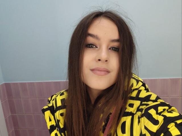 RozmaryKet has long brown hair, light skin, and is wearing a yellow and black hoodie, smiling with brown eyes and pink lips. Her ass is not visible.