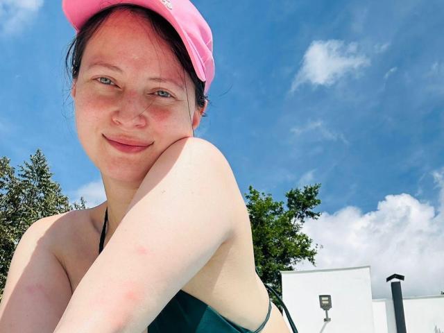 AmbeerWood has fair skin, freckles, and a slight smile, wearing a pink hat and green bikini top, with her arm resting on her cheek. Her tits are partially visible.