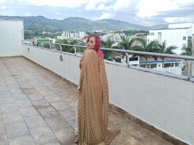 GiselleDunn with pink hair, wearing a brown, wavy-patterned shawl covering her tits and ass, standing on a tiled rooftop, looking back over her shoulder.