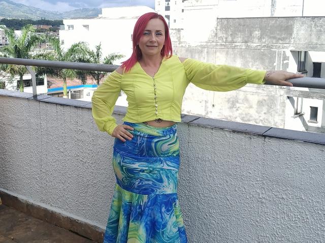 GiselleDunn with pink hair, yellow top, and blue-green swirly skirt, standing on a balcony, right arm on railing, left hand on hip, smiling. Boobs visible under top.