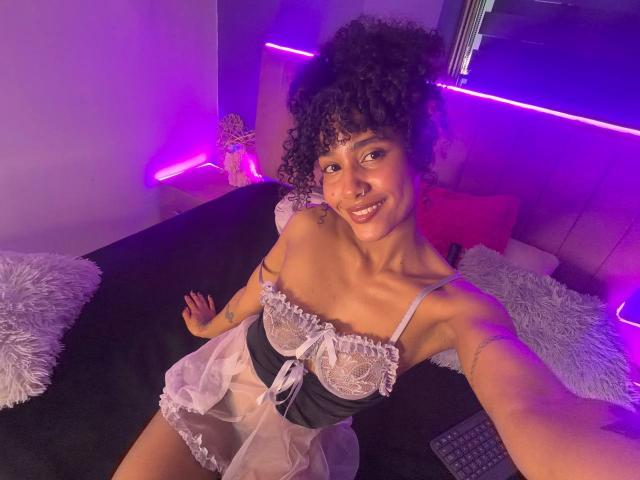 CinthiaWalker with curly hair, smiling, wearing a sheer black and white lingerie dress, sitting on a bed with purple neon lighting, showing her small tits and tattooed arm.