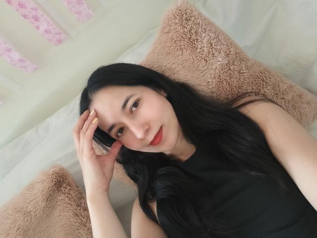 AnneStone with long black hair, red lips, and black sleeveless top, lying on a bed with pink fuzzy pillows, touching her head with one hand, smiling softly.