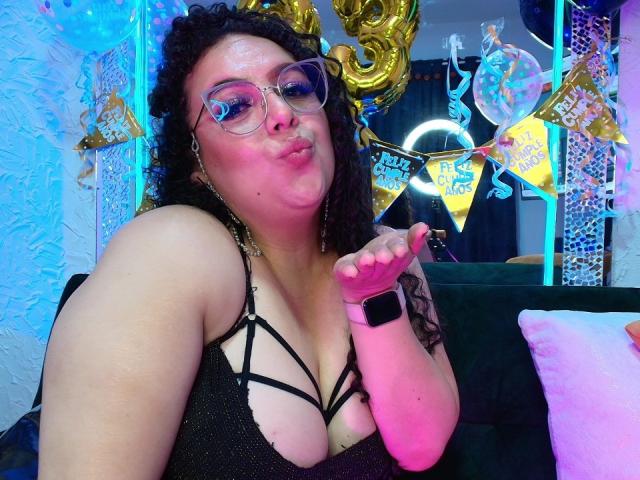 NatashaAmbeer with curly black hair, pink glasses, pouty lips, black strappy top showing her tits, pink watch, gold earrings, and a tattoo on her right arm.