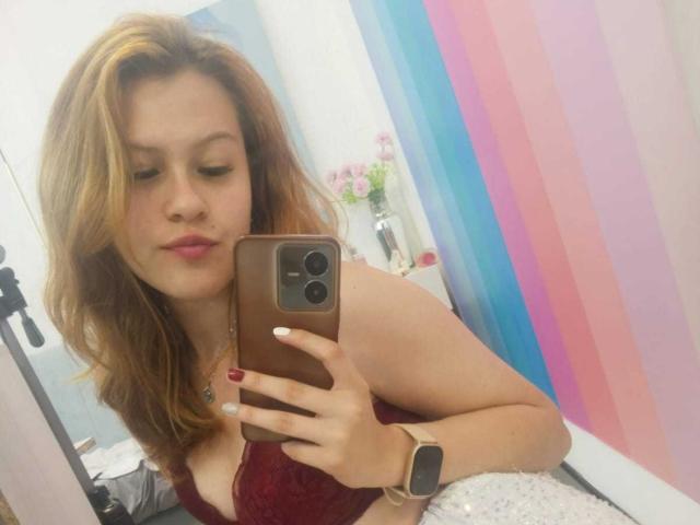 Taylor, usando um sutiã de renda vermelho, segurando um celular, com um relógio no pulso, tem cabelo castanho claro, batom rosa e esmalte vermelho, tirando uma selfie em um quarto com uma parede listrada em tons pastel..