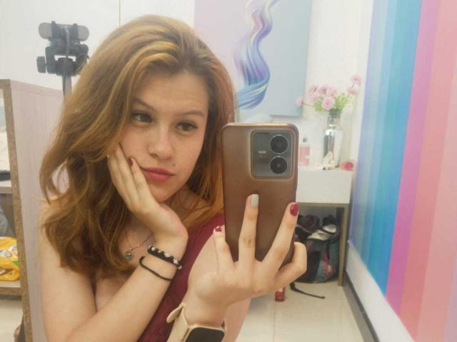 TaylorCarpert with red hair, hand on cheek, holding phone, pink lipstick, red nails, maroon top, mirror selfie, camera in background, striped wall, bracelet, watch.