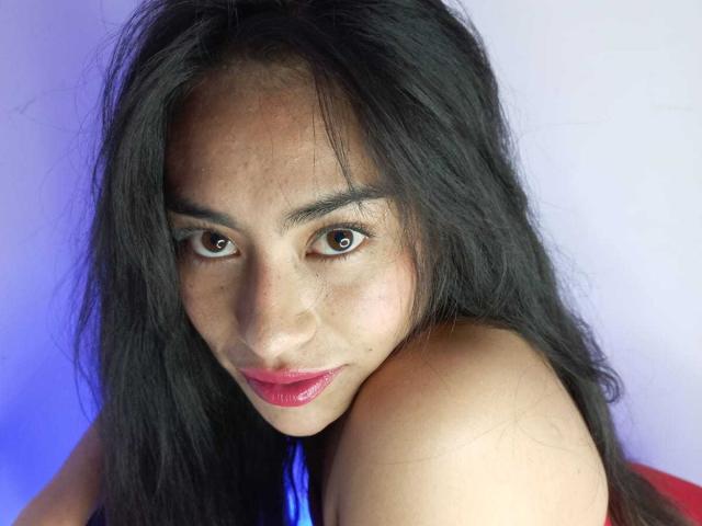 Emily Mitchell tiene el pelo largo y negro, ojos marrones y lápiz labial rojo, y mira directamente a la cámara con una ligera sonrisa, mostrando los hombros desnudos. Es linda y atractiva..