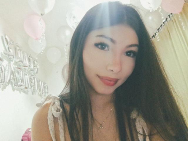 ViolettaLeroy with long brown hair, cute face, and pink lips, wearing a white lace top, smiling softly, light from above, pink and white balloons in background.