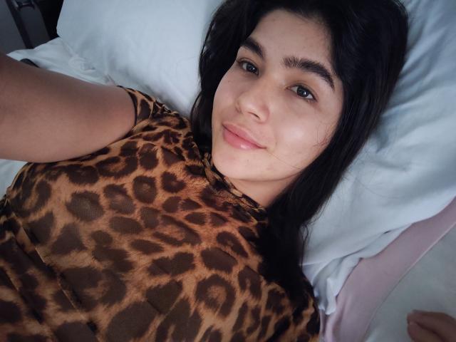 ScarlletZhen with dark hair, brown leopard print shirt, and pink lips, lying on a white pillow, smiling softly at the camera. Her tits are partially visible.
