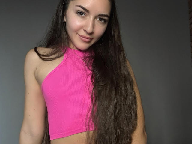 AquaLady with long brown hair, pink ribbed crop top, smiling slightly, showing small tits, fair skin, and brown eyes, against a gray background.