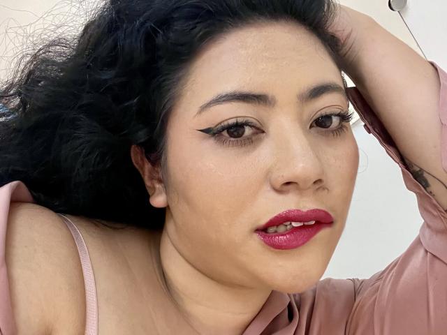 ErikaFox has dark curly hair, brown eyes with winged eyeliner, red lipstick, and is wearing a pink shirt with her hand in her hair. Her ass is partially visible.
