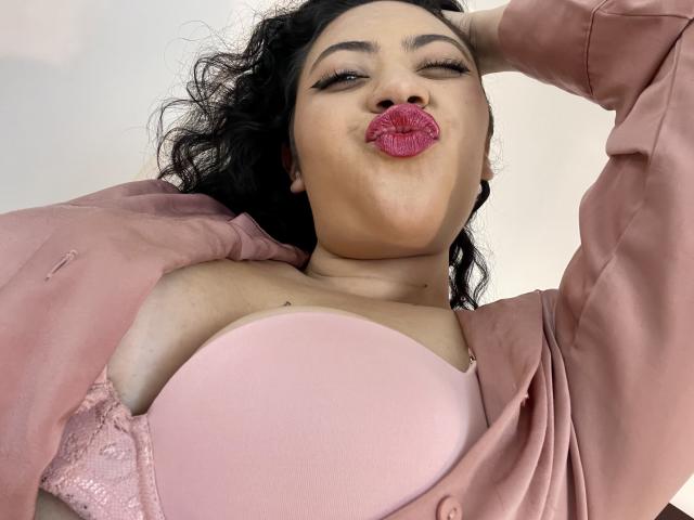 ErikaFox with pink lipstick, pouty kiss, winking, black curly hair, pink satin robe, showing big tits in lace bra, hand in hair, low angle, soft lighting.