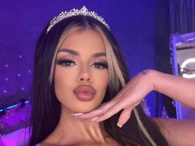 IvyRaven with blonde and black hair, wearing a silver tiara, pouting with full lips, hand near face, blue eyes, long eyelashes, and manicured nails. Purple light background.