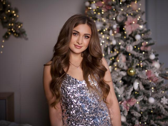 BeataAngel stands in front of a decorated Christmas tree, wearing a sleeveless, silver sequin dress. Her long, wavy brown hair cascades over her shoulders. She has a light smile, brown eyes, and a subtle makeup look. The Christmas tree behind her is adorned with white and pink ornaments, small lights, and a light dusting of snow. The image uses soft, warm lighting to highlight BeataAngel's features and the festive decor.