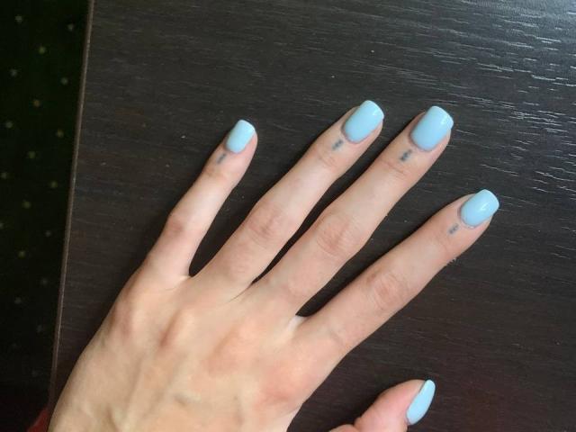 MoonLiza's hand with pale skin, light blue nail polish, and small black tattoos on fingers, resting on dark wooden surface.