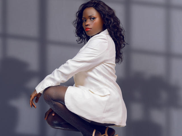 CamiliaWanda with dark skin, curly black hair, wearing a white lab coat, squatting, showing her ass in sparkly black leggings and black heels.