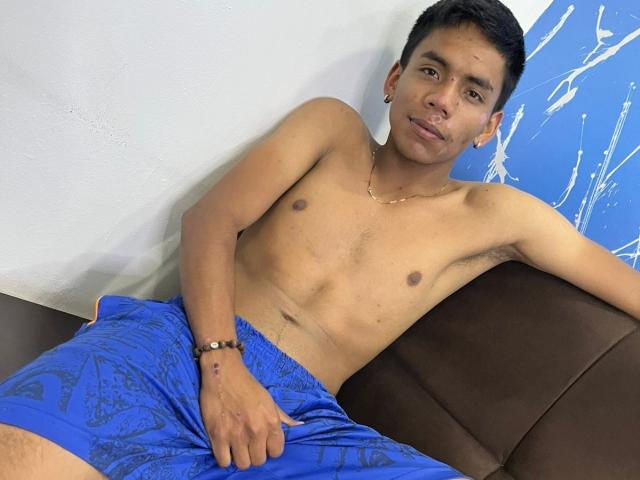 Shirtless Alasttor with short black hair, small brown nipples, and a slight tan, wearing blue patterned shorts, sitting on a brown couch, smiling slightly.