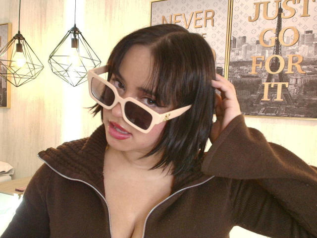 Blueeskyy, con cabello negro hasta los hombros, lleva gafas de sol beige de gran tamaño, una chaqueta marrón con cremallera y lápiz labial rosa, y toca su cabello con una sonrisa juguetona. Sus senos son ligeramente visibles..