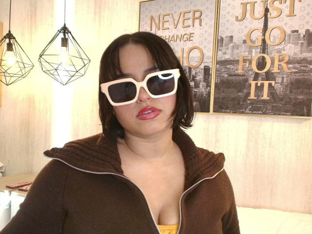 Blueeskyy tiene ojos marrones, lápiz labial rosa, gafas de sol blancas de gran tamaño y una sudadera con cremallera marrón, con una camiseta amarilla debajo, que deja ver unos pechos pequeños. Tiene un rostro lindo y curvilíneo..