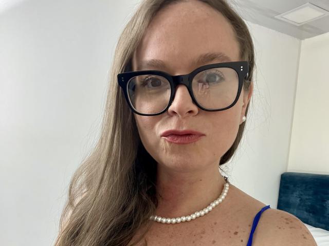 PamelaColin has glasses, a pearl necklace, and brown hair, with freckles on her face, wearing a blue top, and has a serious expression, with a slight pout.