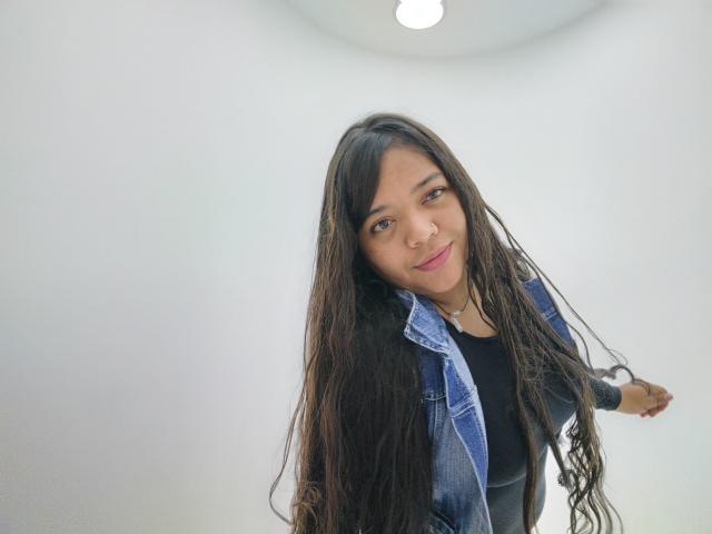 Shienna with long, wavy dark hair, wearing a blue denim jacket over a black top, smiling slightly, brown eyes, light brown skin, small nose, and pink lipstick.