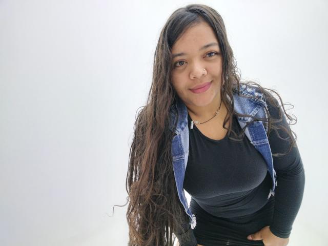 Shienna with long, wavy brown hair, wearing a denim vest over a black shirt, smiling slightly, hands by her sides, light skin, brown eyes, and pink lipstick.