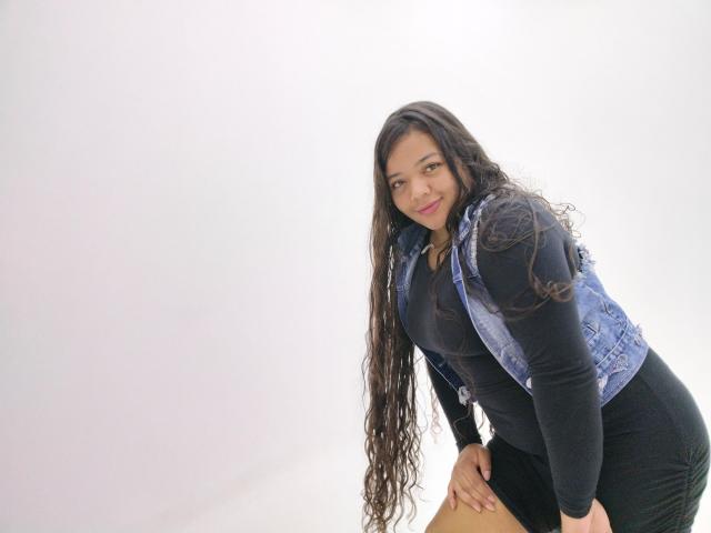 Shienna with long, wavy dark hair, wearing a denim vest over a black long-sleeve top and black skirt, leaning forward with a slight smile, showing her ass.