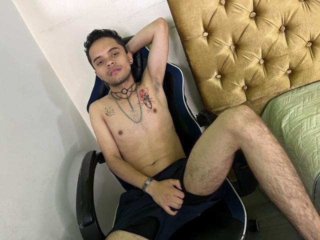 MinhoReyes, shirtless, with tattoos, dark hair, wearing black shorts, sitting in chair, arm behind head, hand on thigh, gold tufted headboard, green quilted bed, relaxed, confident expression.