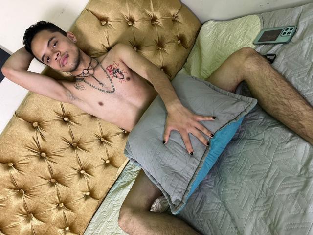 MinhoReyes, shirtless with tattoos, lying on a gold-tufted bed, holding a gray pillow, legs spread, phone on bed, black nail polish, looking at camera, slightly pouty face.