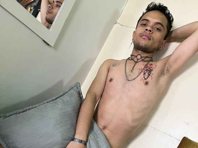 Shirtless MinhoReyes with tattoos, black hair, and a chain necklace, leans against a wall with a relaxed, confident expression. He's wearing grey pants, and there's a mirror reflection.
