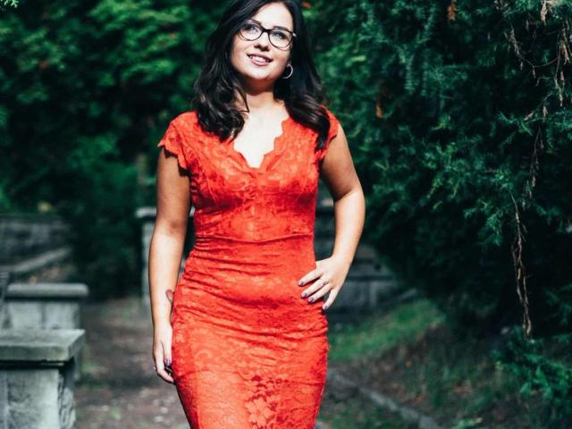 CuteLexy-hot in a red lace dress, standing with a hand on her hip, has glasses, dark wavy hair, and a smile. Her tits are medium, her ass is curvy.