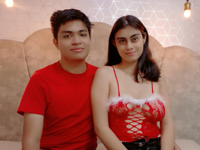 SofiNdLogan in a red shirt, sitting next to SofiNdLogan in a red lace-up lingerie top with white fur trim, on a beige couch. Her tits are visible.