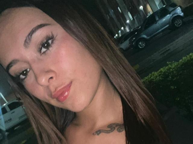 DamiAmbani with glossy pink lips, long brown hair, and a nose ring, showing off her small tits and a tattoo just below her collarbone in a night selfie.