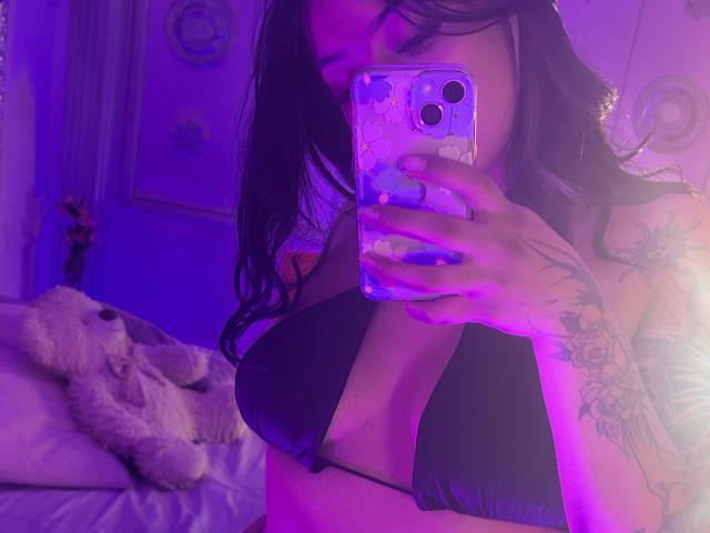 KatianPierce with dark hair, black bikini, tattoos on right arm, holding phone, purple and blue lighting, visible breasts, taking mirror selfie, fluffy stuffed toy on bed.