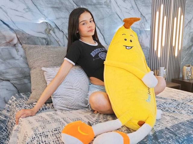 MiaGalvez with yellow banana pillow, wearing black shirt, blue shorts, sitting on bed, gray pillow, blue patterned bedspread, long dark hair, looking at camera, relaxed, cute.