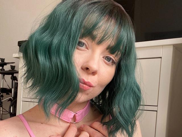 JessieFire-hot with teal green wavy hair, pink collar, pouty pink lips, and gray eyes, wearing a pink top, hands near neck, in a soft-lit room.