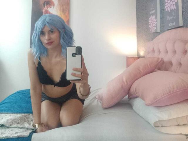 LilaaSalmon with blue wig, black lace bra, and black panties, kneeling on bed, holding phone, showing her tits and ass, pink pillows, white walls, and blue blanket.