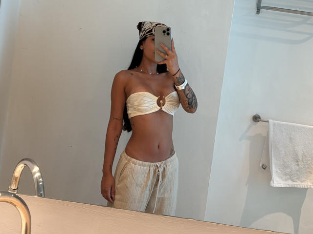 ThaliaAres in a mirror selfie, wearing a white bandeau with a gold ring, loose pants, and a patterned headscarf, showing her tats, tan skin, and small tits.