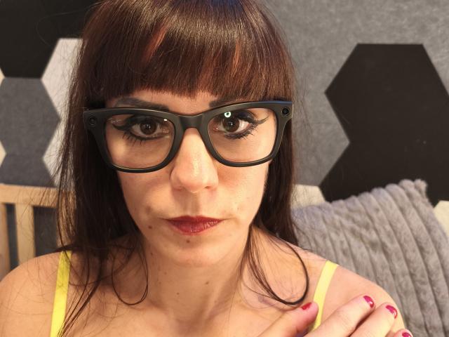 OrianaLaFrancaise with brown hair, black glasses, red lipstick, yellow bra, and pink nails, showing off her face and slight pimple on her left cheek.