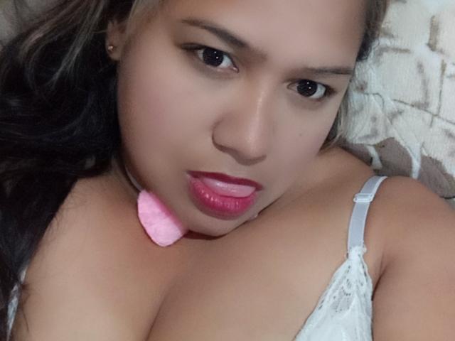 CandyCurvy hat eine hellbraune Haut, pinken Lippenstift und eine pinke Herz-Kette. Sie schaut mit leicht zusammengepressten Lippen in die Kamera und trägt einen weißen Spitzen-BH. Brüste sind sichtbar..