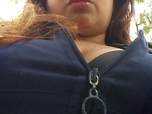 CandyCurvy's face is partially visible with her lips slightly pouty. She's wearing a black zip-up, exposing her black bra and showing her large, round tits.