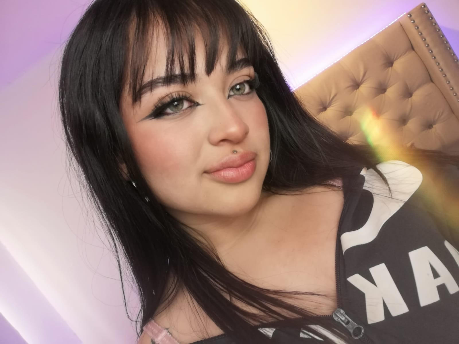 KokoLette has black hair, green eyes, and a nose ring. She's wearing a black and white top, pouting with pink lips, and looking at the camera.