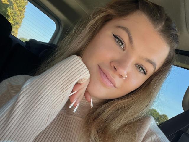 Avaexklusiv-hot with long brown hair, green eyes, and a nose ring, wearing a beige ribbed sweater, smiling, head on hand, with white nails, in a car.